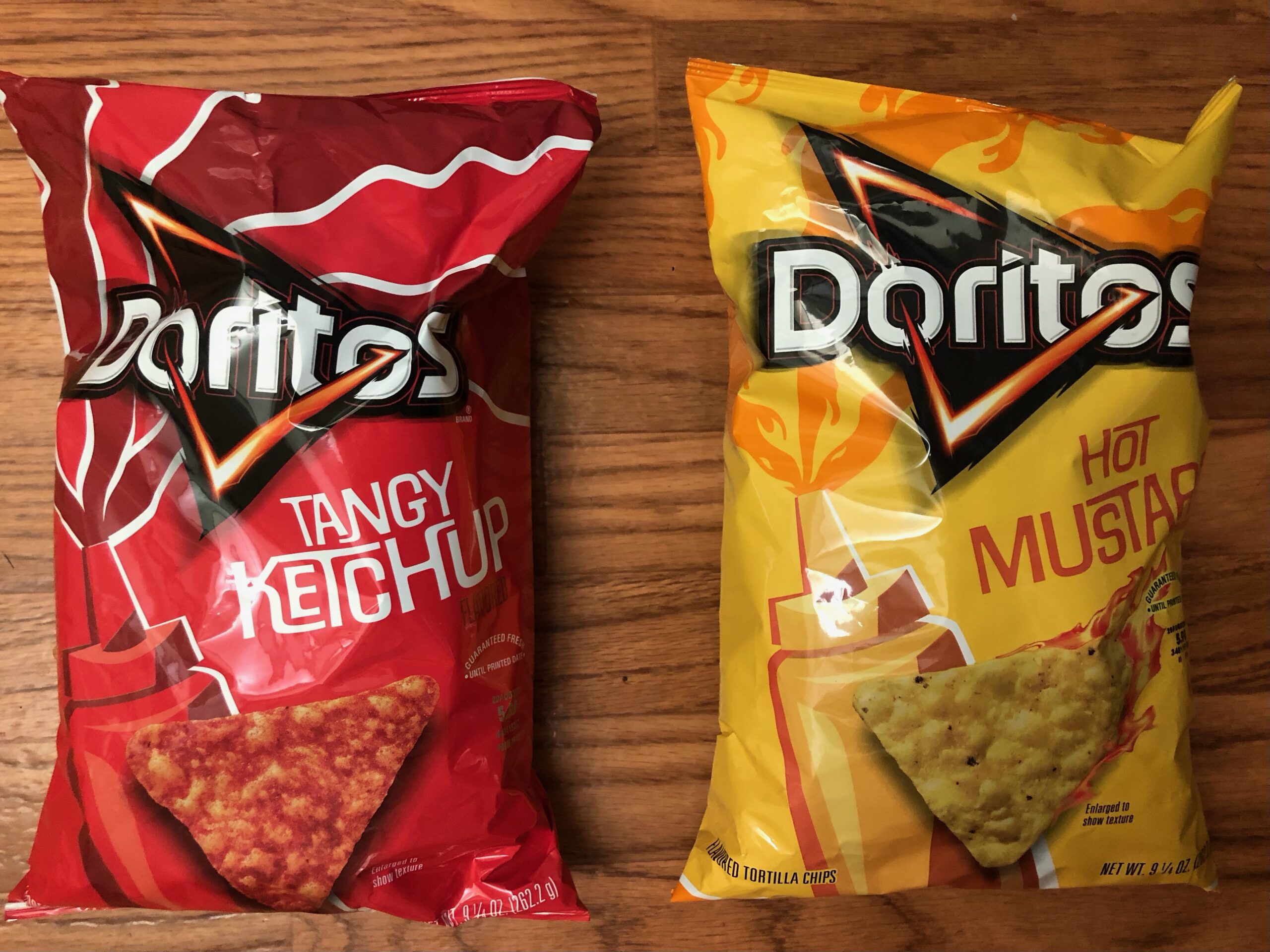 A red bag of Doritos Tangy Ketchup on the left and a yellow bag of Doritos Hot Mustard on the left. Both bags are laid upon a wooden background.