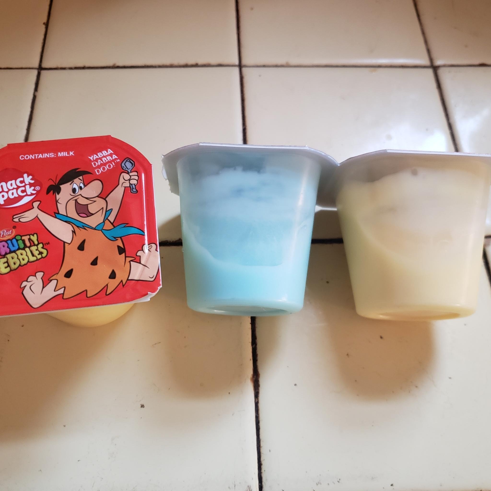 Three pudding cups on a beige tiled counter. The one on the right is upright and shows Fred Flintstone jumping with a spoon. The two on the left are on their sides. One is filled with light blue pudding and one is filled with yellow pudding.