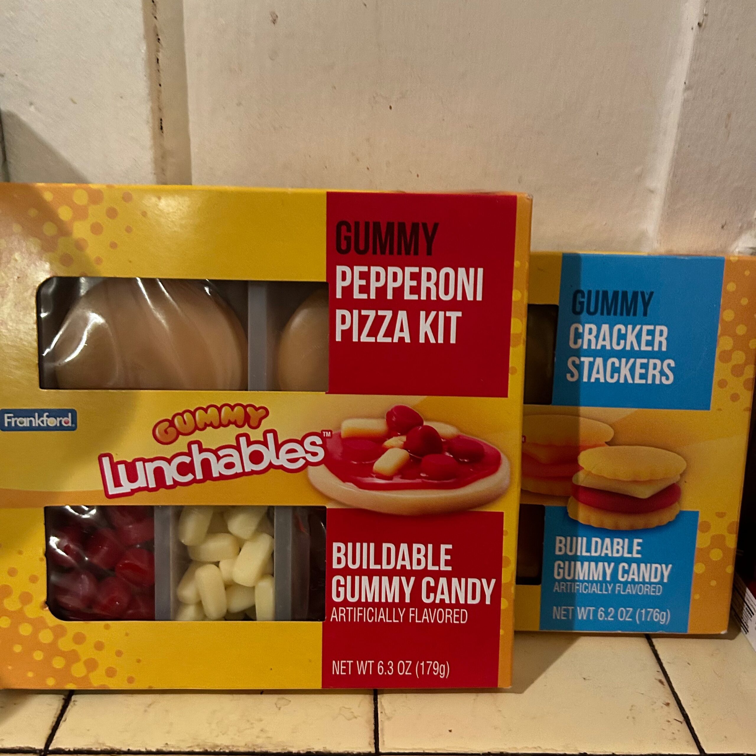 Two boxes of "gummy lunchables." One is a pizza kit and the other is cracker stackers. The two boxes are posed on a yellow countertop with a white wall.