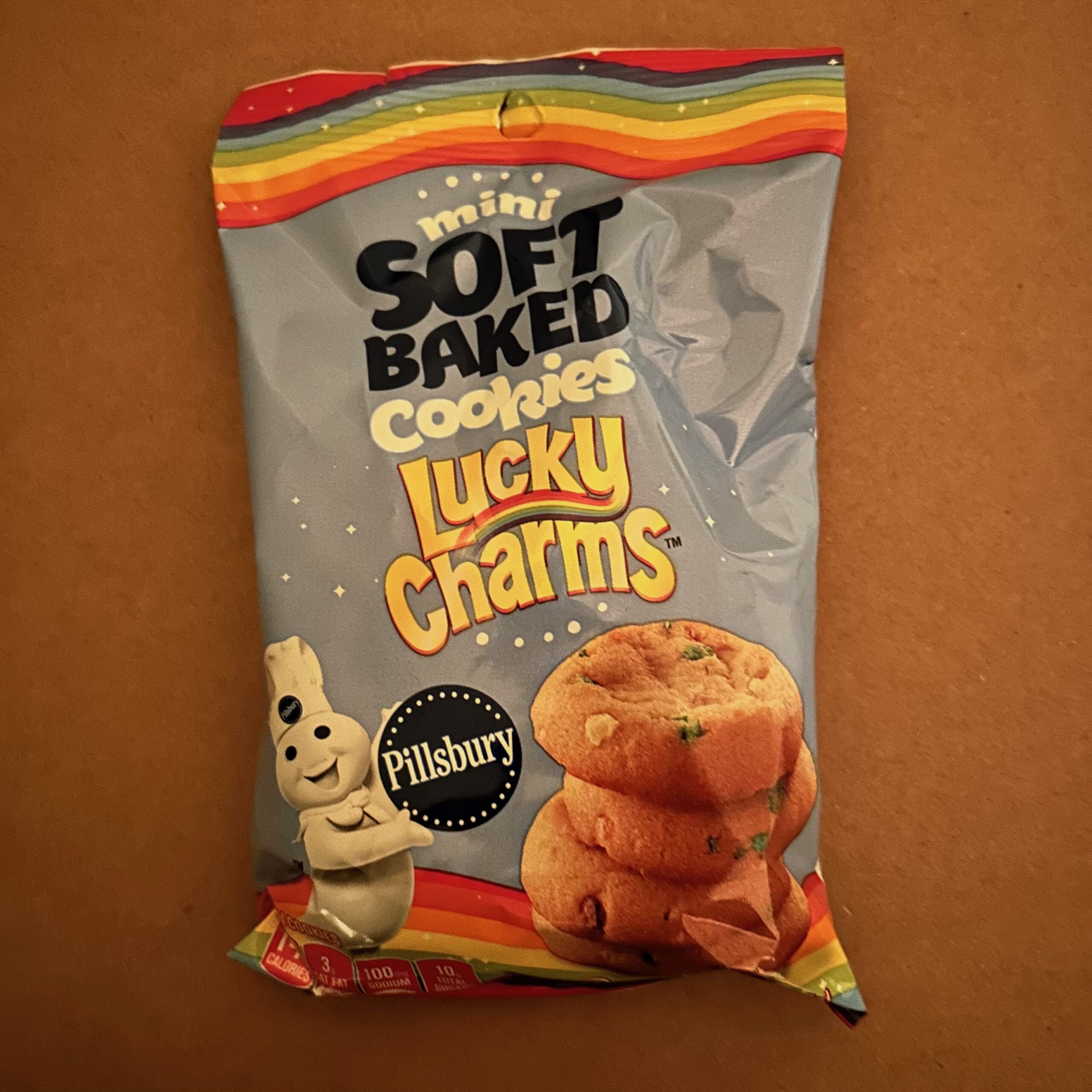 A dim brown background and and a bag which reads "soft baked cookies lucky charms." There is an image of thick cookies in the lower right of the bag, the Pillsbury mascot holding the Pillsbury logo in the bottom left, and a rainbow stripe across the top of the bag.