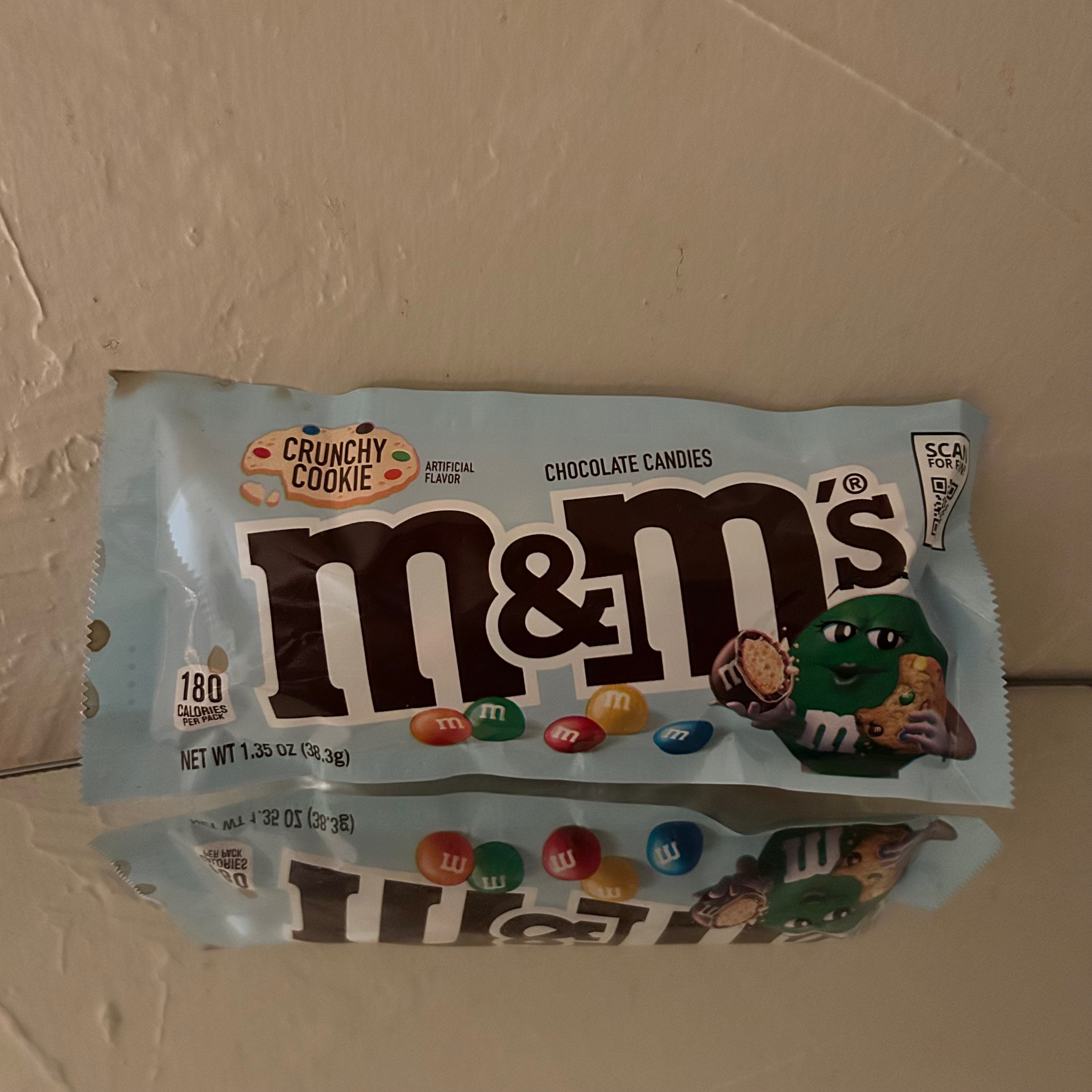 A beige wall in the background and a mirrored surface underneath a blue bag of crunchy cookie M&M's. The bag features the green M&M mascot holding half of a brown M&M in one hand and a bitten-into cookie in the other hand.